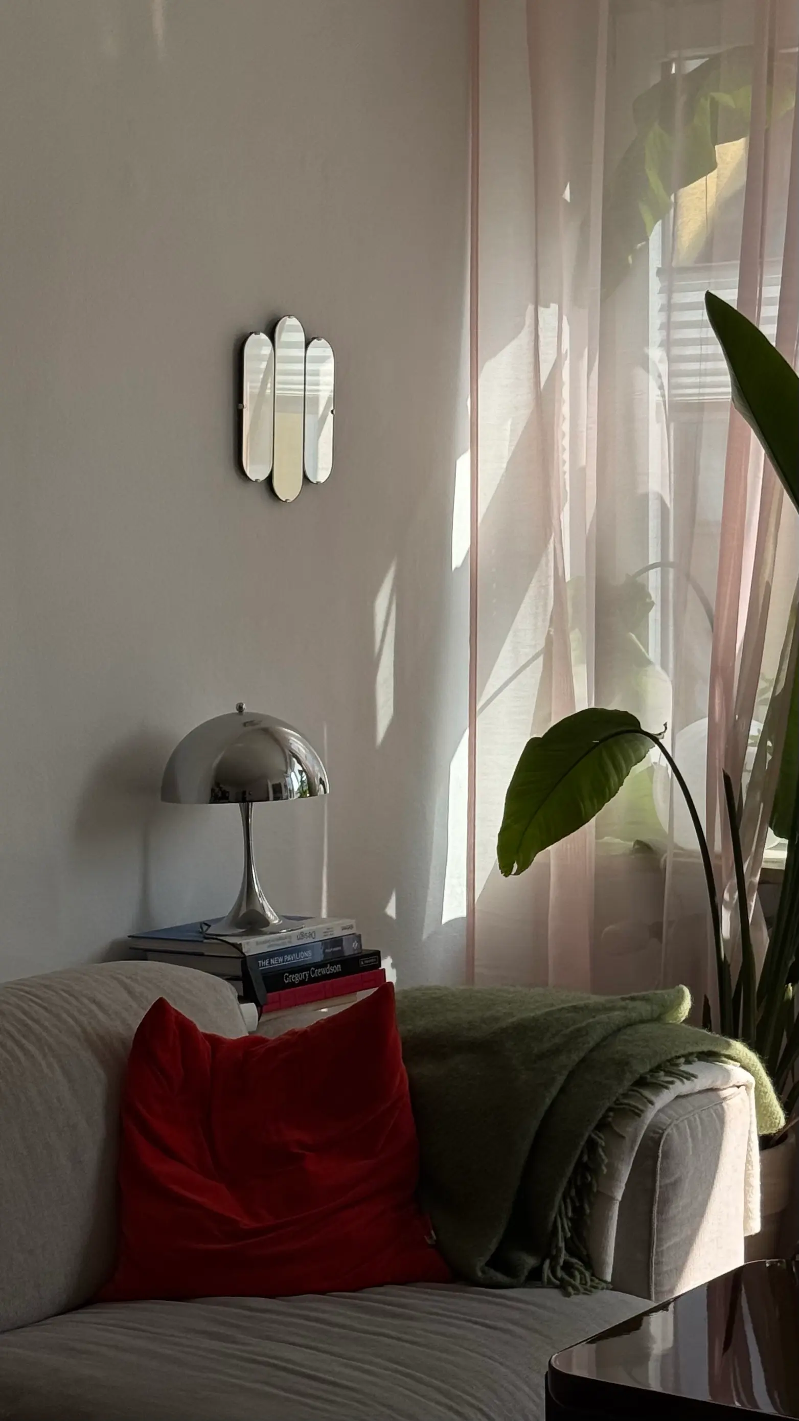Wohnzimmerecke im Nachmittagslicht: Sofa mit rotem Samtkissen und olivgrüner Decke, Chrom-Pilzlampe auf einem Bücherstapel, Vorhang und Strelitzie am Fenster